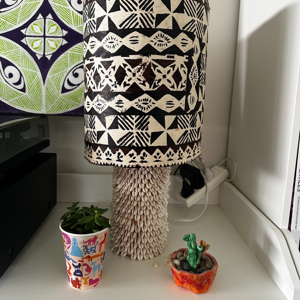 A beautifully repurposed Kmart lamp decorated with hand-glued shells and wrapped in Fijian masi cloth. This handmade light adds a gentle, coastal glow to the calm corner, blending creativity, culture, and family craftsmanship in one cosy space.