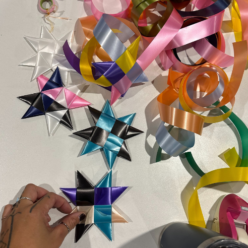 Hand weaving colourful ribbon stars on a white table with pink, blue, black and yellow ribbons scattered around — promoting mindfulness and creative calm for World Mental Health Day.