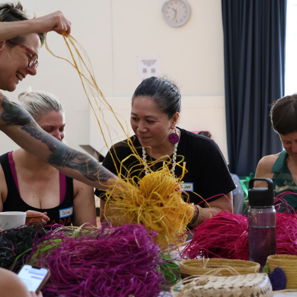 Women sitting and weaving with coloured raffia on the table.